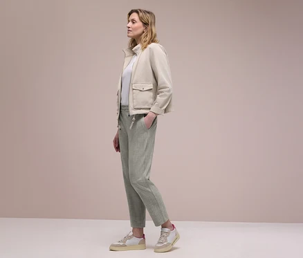 Frau im Profil trägt eine beigefarbene Jacke, ein weißes Oberteil und hellgrüne Street One Joggpants, eine Hand in der Tasche. Sie trägt beige-weiße Sneaker.