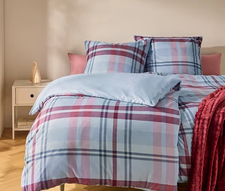 Ein Bett mit einer blau, rosa und rot karierten Bettdecke und Kissen. Ein rotes Plaid liegt auf der rechten Seite. Links steht ein Nachttisch mit einer Lampe.