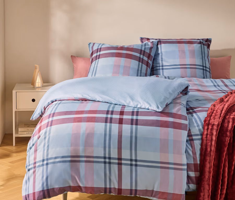 Ein Bett mit einer blau, rosa und rot karierten Bettdecke und Kissen. Ein rotes Plaid liegt auf der rechten Seite. Links steht ein Nachttisch mit einer Lampe.