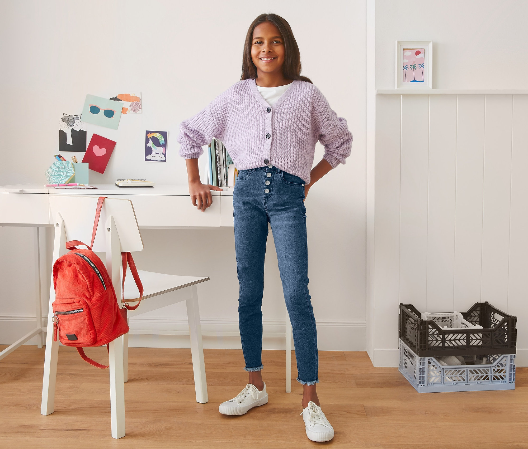 Lächelndes junges Mädchen posiert in blauen High-Waist-Jeans und lila Strickjacke vor einem Schreibtisch.