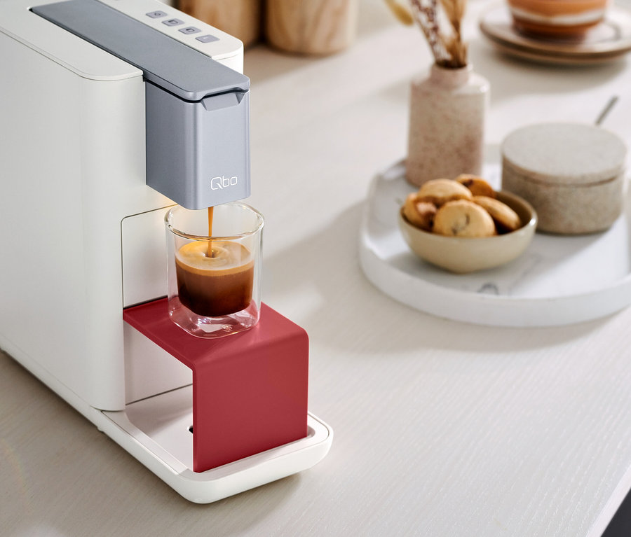 Qbo ESSENTIAL Bright Coral Kaffeemaschine befüllt eine Tasse mit Kaffee. Kekse stehen auf dem Tisch.