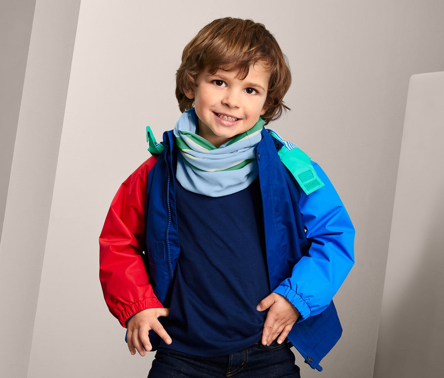 Ein Junge mit braunen Haaren lächelt und trägt ein blau-grünes, recyceltes Multifunktionstuch, ein blaues T-Shirt und eine farbige Jacke.