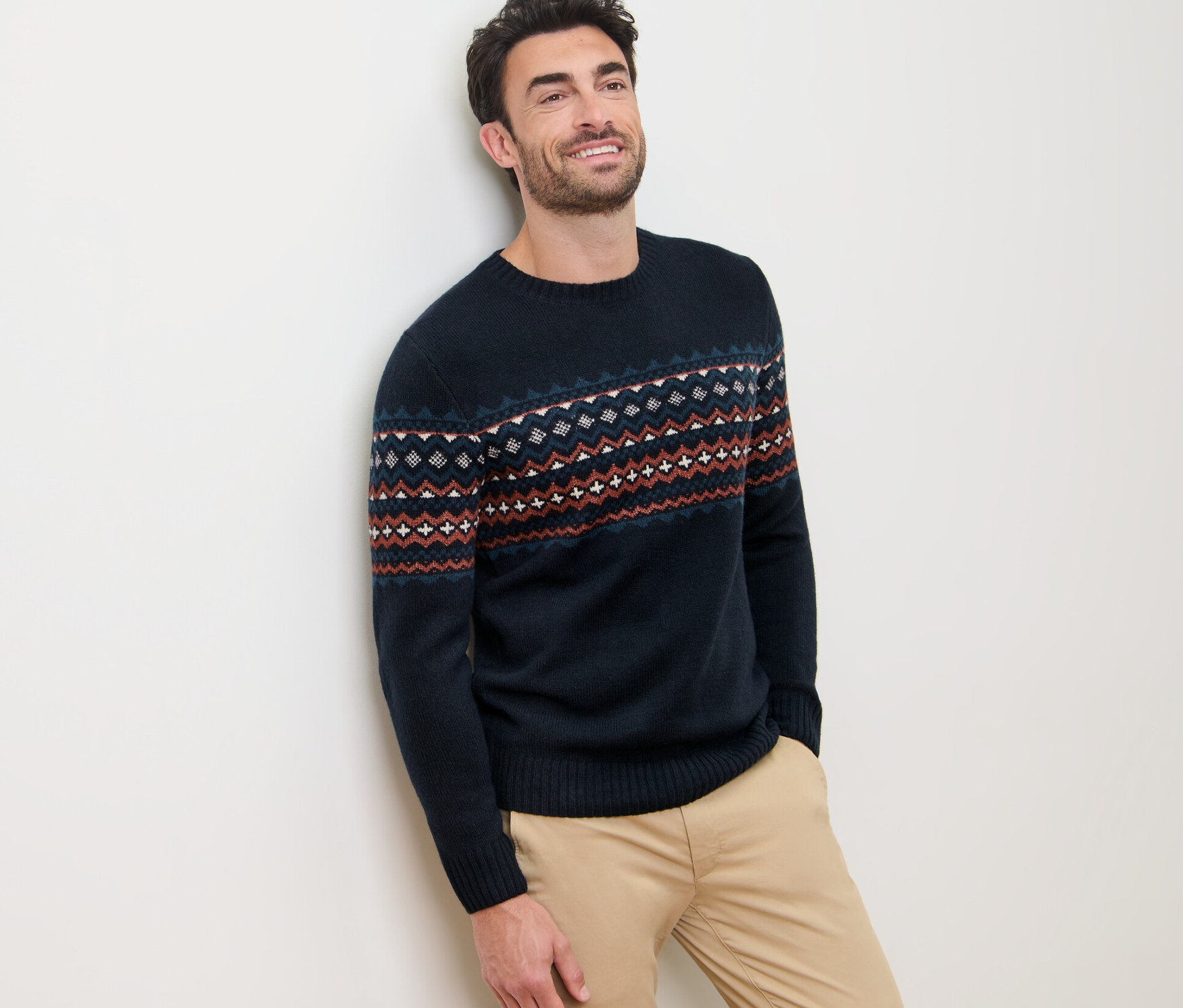 Mann lehnt an Wand, trägt Strickpullover.