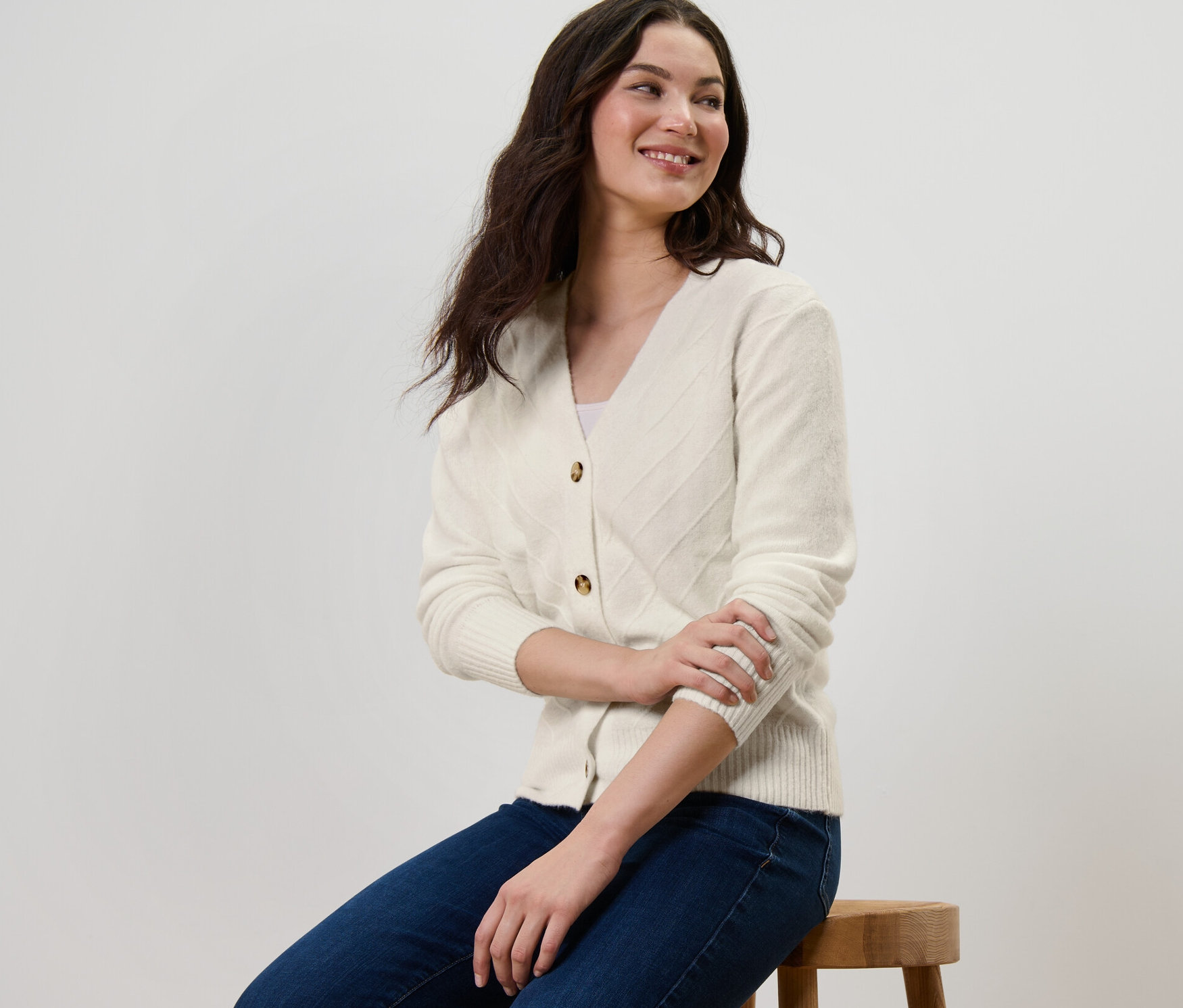 Eine Frau sitzt auf einem Hocker und trägt einen cremeweißen Cardigan mit Strickstruktur.