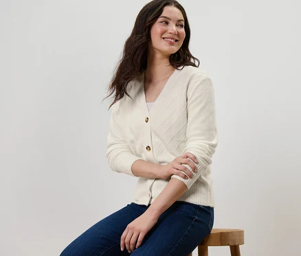 Eine Frau sitzt auf einem Hocker und trägt einen cremeweißen Cardigan mit Strickstruktur.