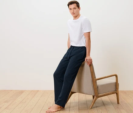 Mann in weißem T-Shirt und Relaxhose lehnt an beigem Sessel.