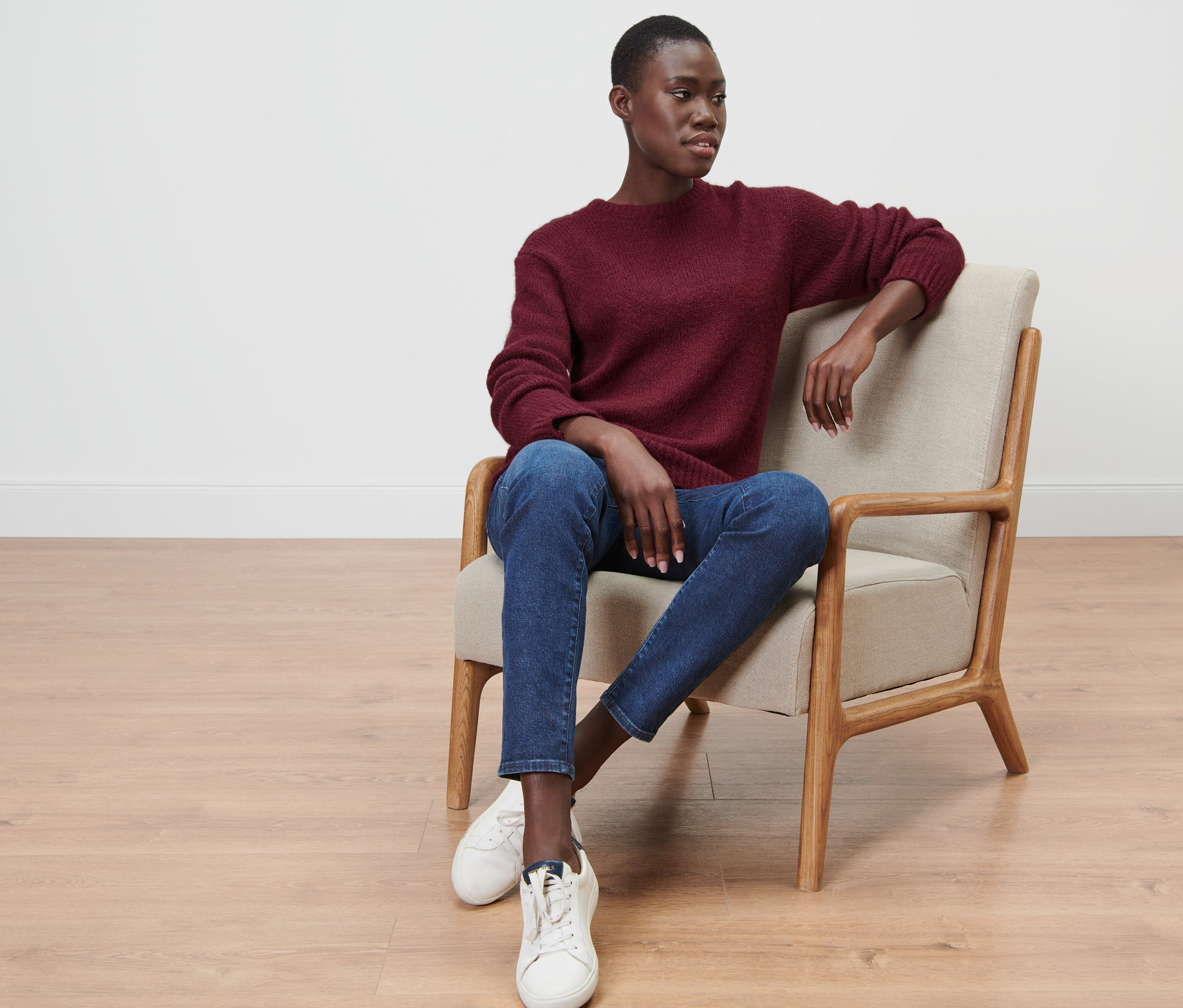 Frau sitzt in Sessel, trägt bordeauxroten Grobstrickpullover mit Wolle, Jeans und Turnschuhe.