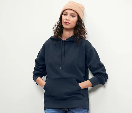 Frau mit braunen lockigen Haaren und Mütze posiert in dunkelblauem Hoodie.