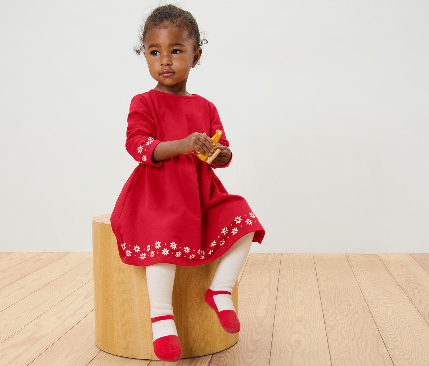Ein Mädchen sitzt auf einem Hocker und trägt ein rotes Kinder-Sweatkleid mit weißen Blumen.