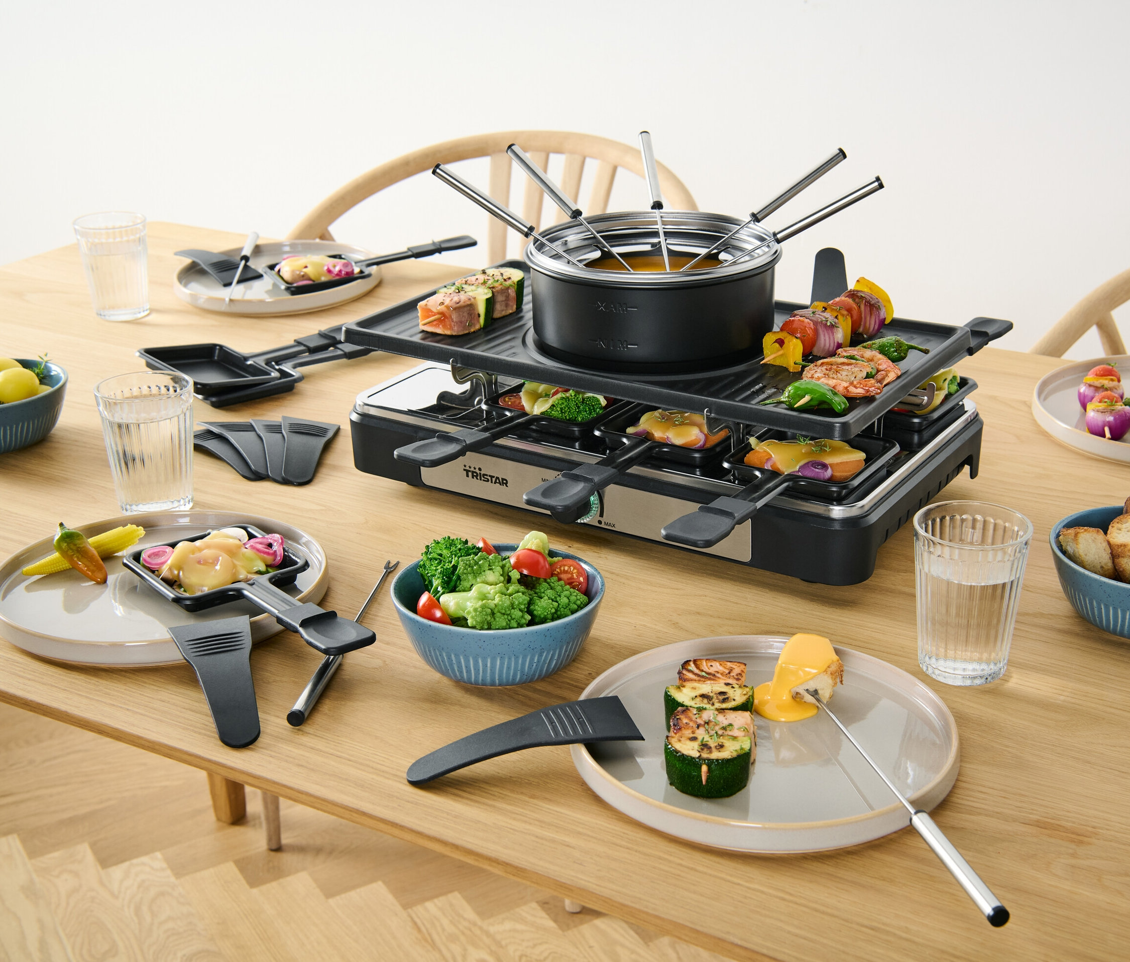 Ein 2-in-1-Raclette-und-Fondue-Gerät steht auf einem Tisch mit Speisen.