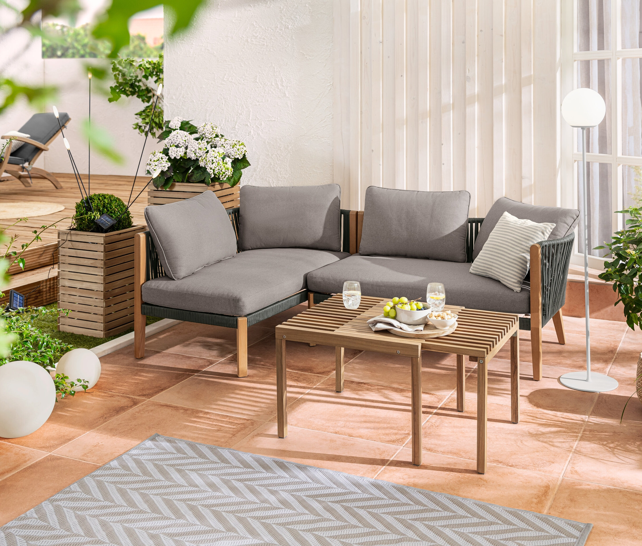 Lounge-Ecke mit Textilgeflecht auf einer Terrasse. Dazu Solar-Kugelleuchten (20 und 30 cm), LED-Akku-Stehleuchte und ein Pflanzsäulen-Set »Tinus«.