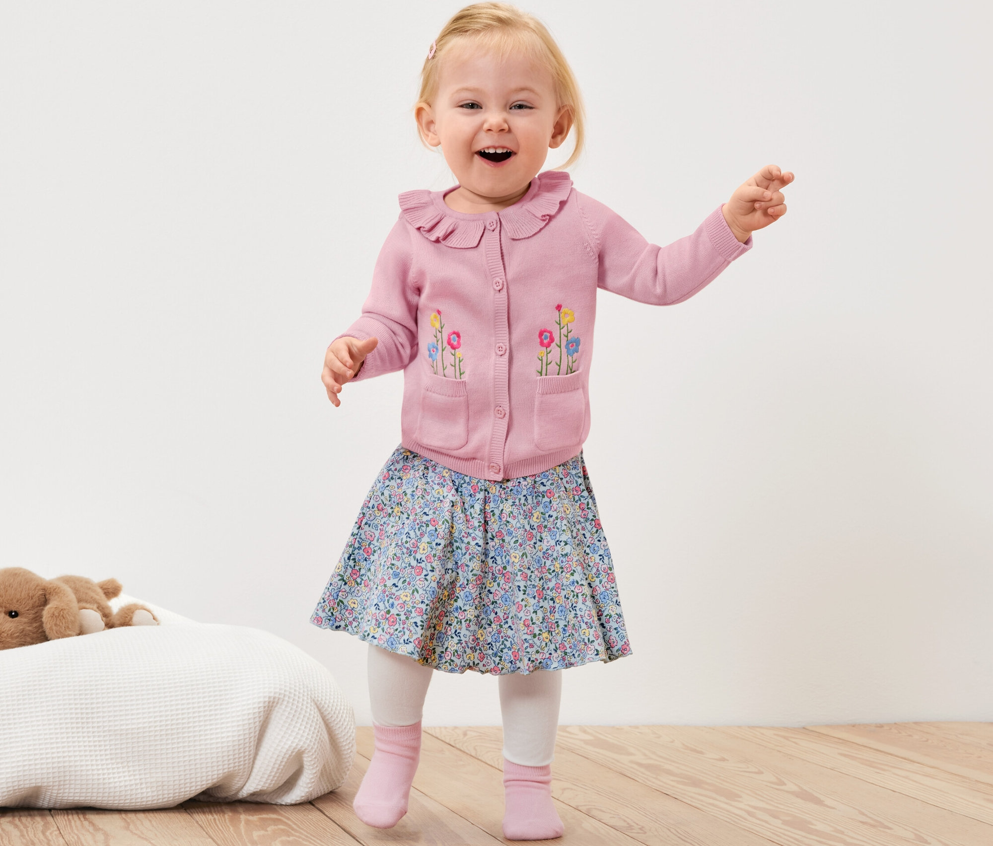 Lächelndes Kleinkind winkt mit den Händen und trägt eine rosa Baby-Strickjacke mit Blumen, einen geblümten Rock und rosa Socken.