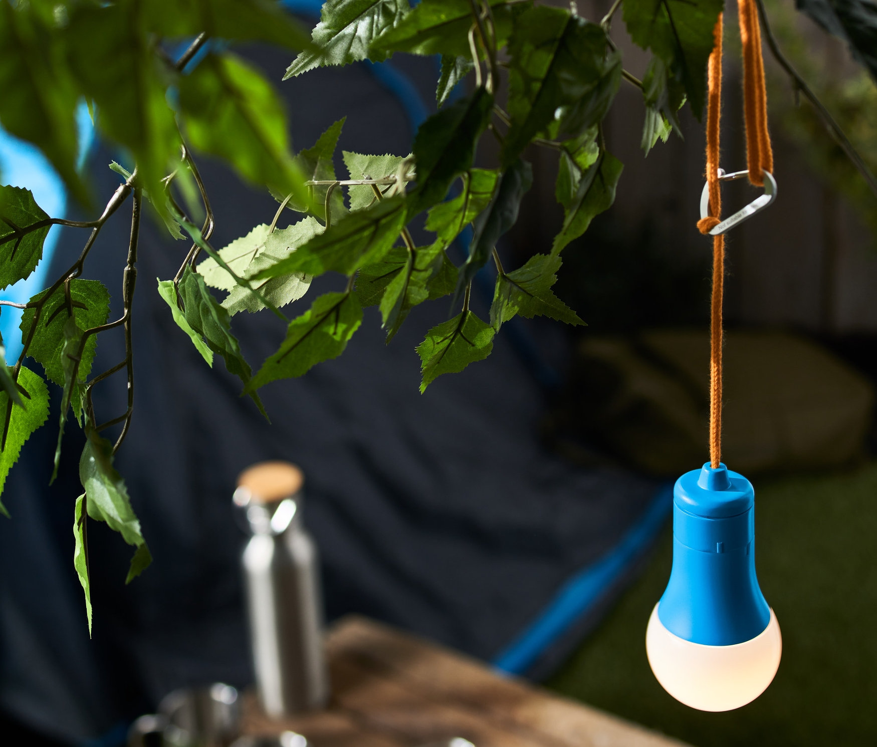 Eine blaue LED-Camping-Lampe hängt an einem Ast, im Hintergrund ein Zelt.