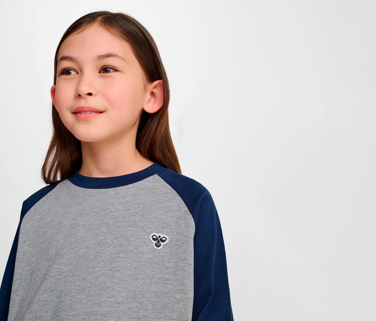 Porträt eines jungen Mädchens, das ein graues HMLJR Raglan Loose T-Shirt mit blauen Ärmeln trägt.