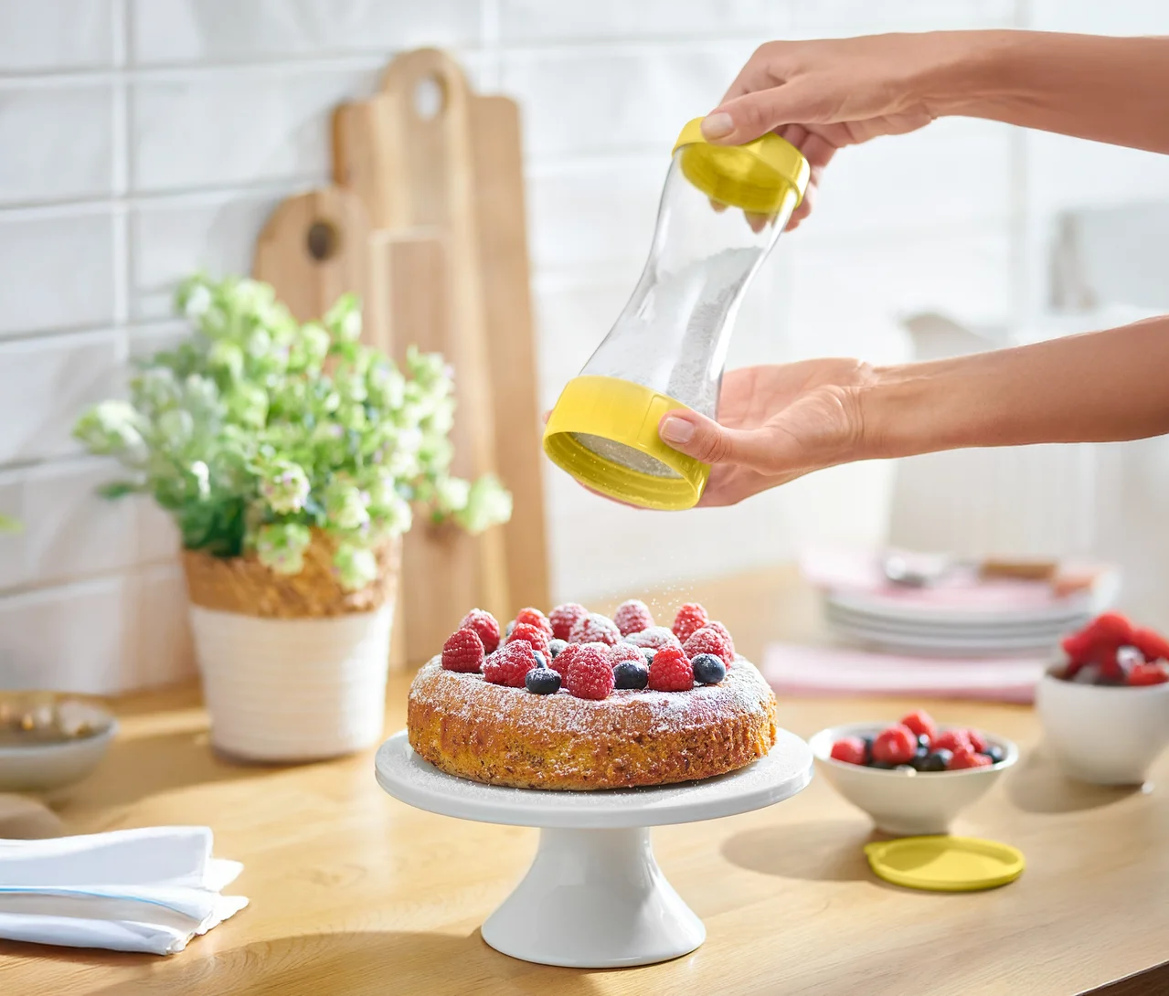 Eine Person streut Puderzucker mit einem gelben Streuer auf einen Kuchen mit Beeren.