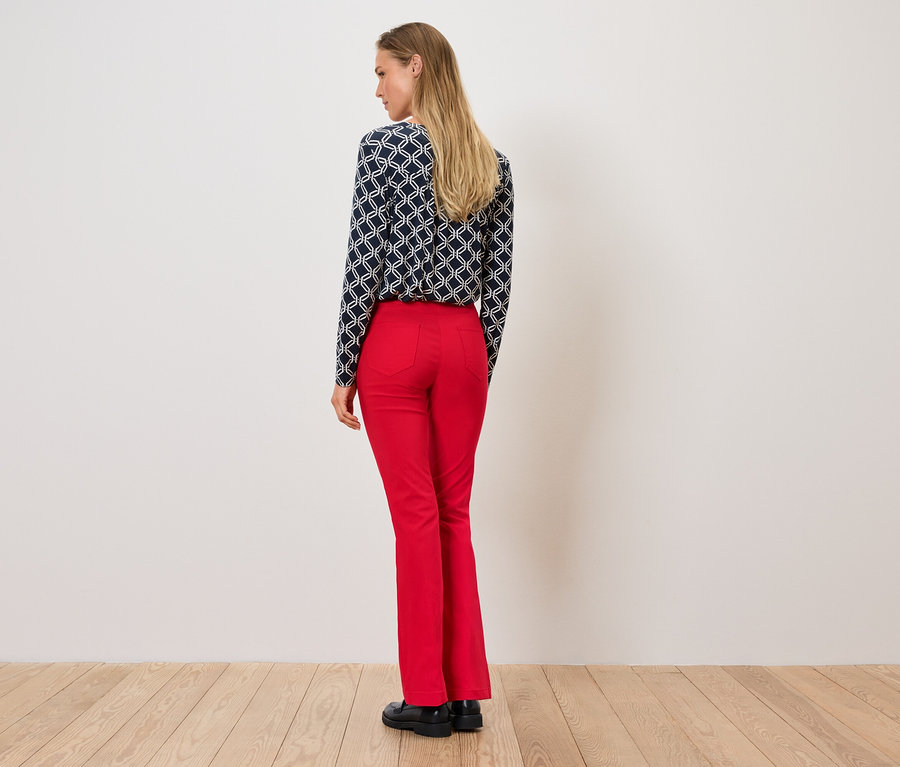 Frau steht mit dem Rücken zur Kamera, trägt eine gemusterte Bluse und rote Hose.