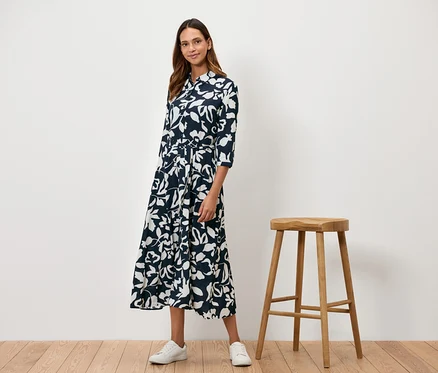Eine Frau steht neben einem Holzhocker und trägt ein marineblaues Kleid mit weißem Blumenmuster.