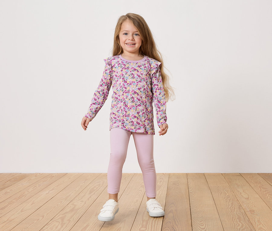 Ein Mädchen mit blonden Haaren trägt ein geblümtes Hemd und rosa Leggings und weiße Schuhe.