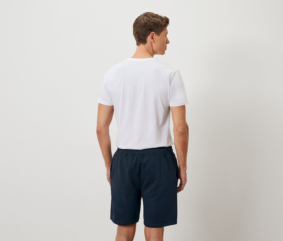 Mann von hinten in einem weißen T-Shirt und marineblauen Shorts vor einem neutralen Hintergrund.