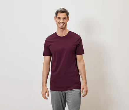 Mann mit grau melierten Haaren lächelt in einem burgunderroten T-Shirt und grauer Hose vor einem weißen Hintergrund.