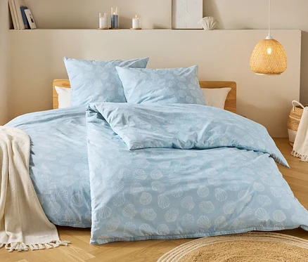 Ein Schlafzimmer zeigt ein hellblaues Bettzeug mit Muschelmuster, einen Holzhocker mit Decke und eine geflochtene Lampe.