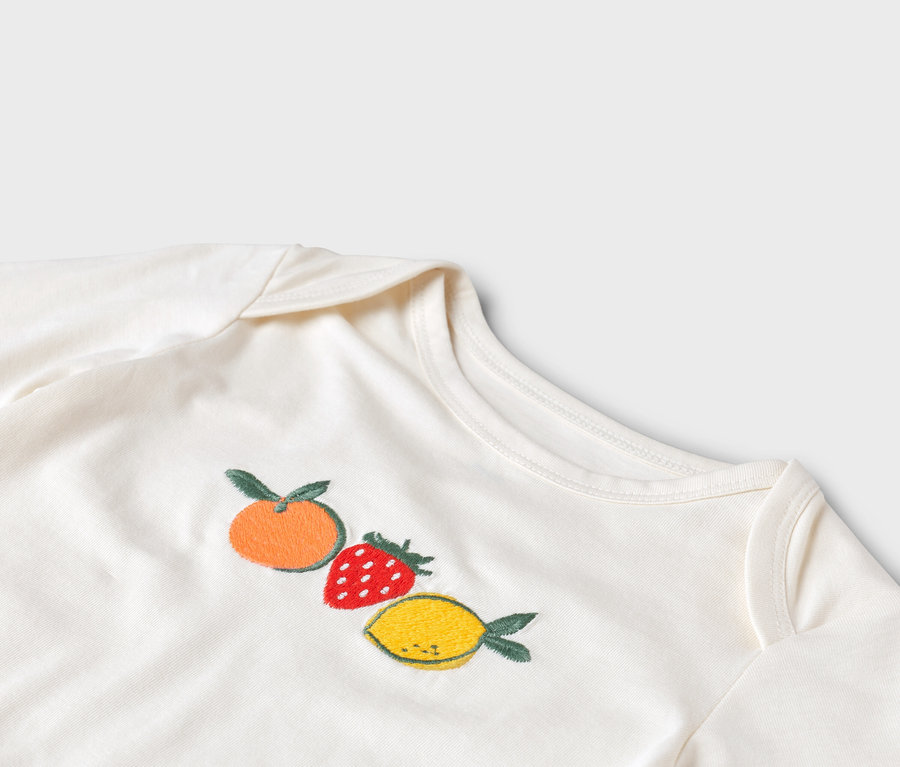 Nahaufnahme eines weißen Baby-Bodys mit einer gestickten Mandarine, Erdbeere und Zitrone.