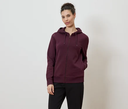 Frau mit Dutt trägt eine weinrote Kapuzenjacke mit Reißverschluss und schwarze Hose vor einem weißen Hintergrund.