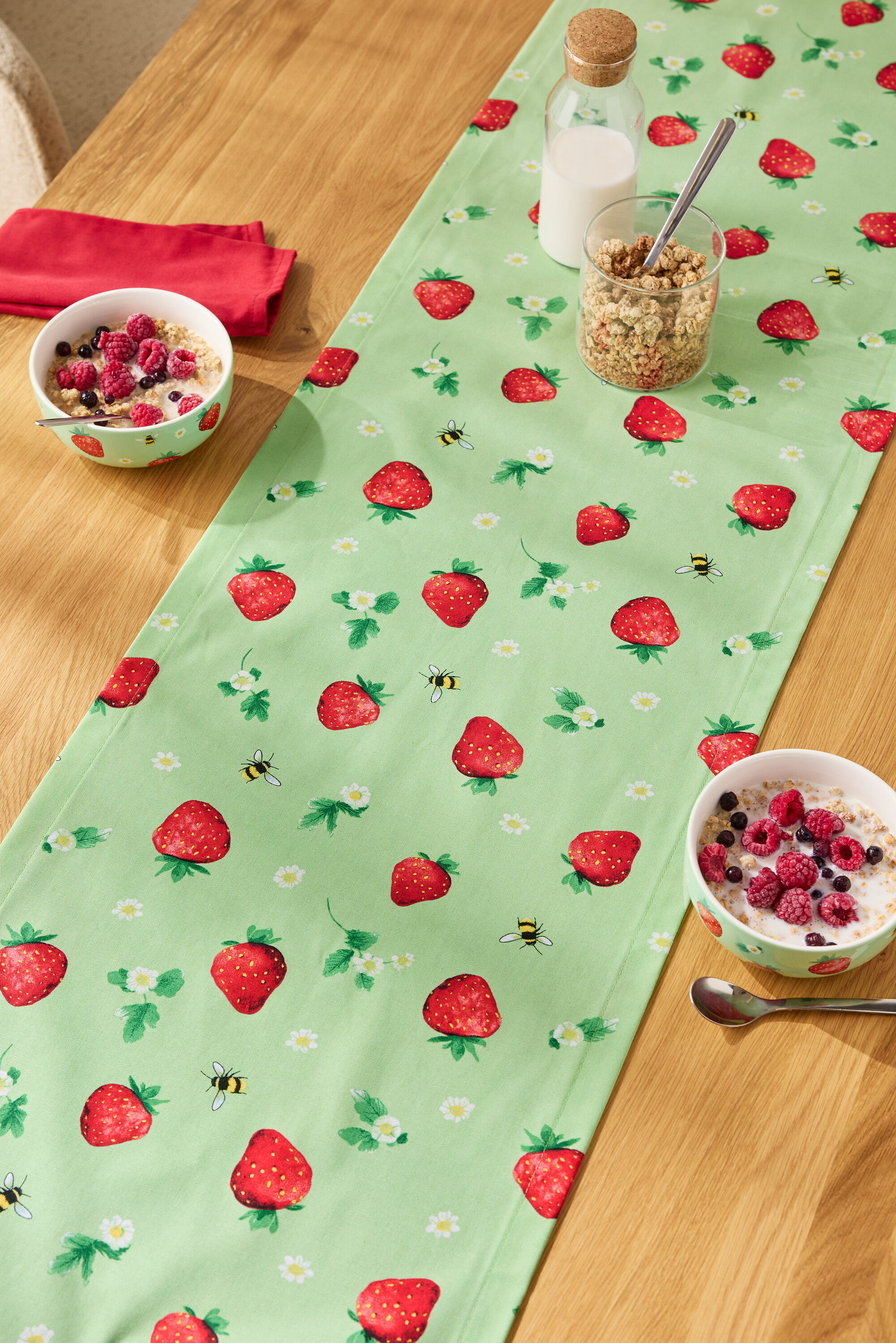 Auf einem Holztisch liegt ein Tischläufer mit Erdbeermuster, darauf Müslischalen, ein Glas Müsli und eine Milchflasche.