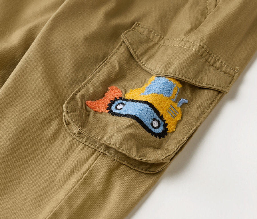 Nahaufnahme einer khakifarbenen Cargohose mit aufgesetzter Tasche, die mit einem gestickten Bagger in Orange, Gelb und Blau verziert ist.