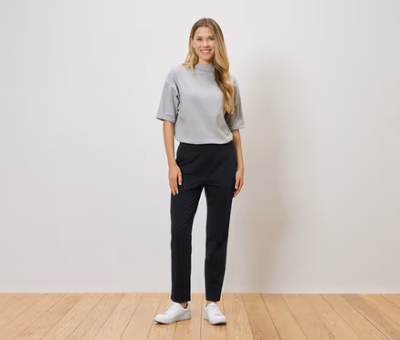 Eine Frau mit blonden Haaren steht vor einer weißen Wand und trägt ein graues T-Shirt, eine schwarze Hose und weiße Turnschuhe.
