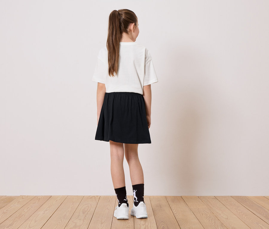 Rückansicht eines jungen Mädchens mit Pferdeschwanz, weißem T-Shirt, schwarzem Rock und Socken mit einer weißen Figur, die auf einem Holzboden steht.