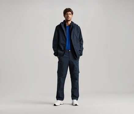 Ein Mann steht mit seinen Händen in den Taschen einer dunkelblauen Jacke und einer passenden Hose, dazu trägt er ein blaues T-Shirt und weiße Turnschuhe.