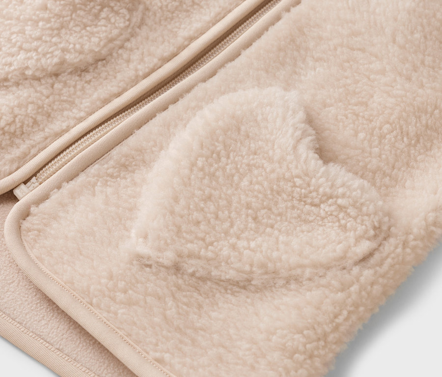 Nahaufnahme einer cremefarbenen Teddyjacke mit Reißverschluss und einer herzförmigen Tasche.