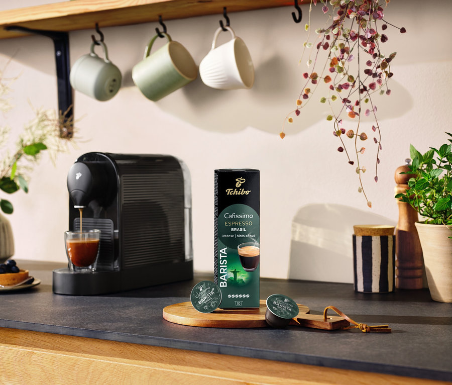 Schwarze Kaffeemaschine bereitet Kaffee in einem Glas neben einer Packung Cafissimo BARISTA Espresso Brasil - 10 Kapseln zu.