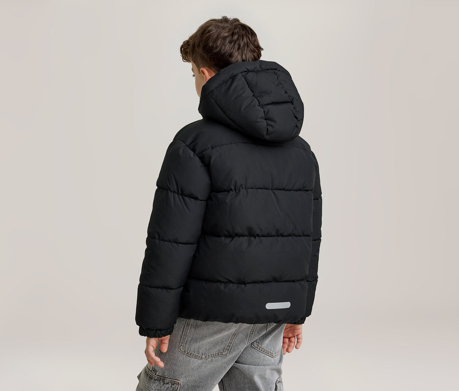 Ein junger Junge von hinten, der eine schwarze Kapuzen-Steppjacke und graue Jeans trägt. Die Jacke hat horizontale Steppnähte und einen reflektierenden Streifen am unteren Rücken.