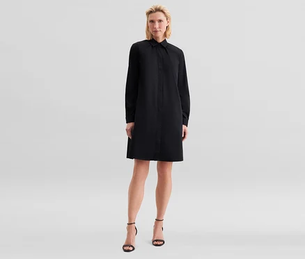 Blonde Frau steht in einem schwarzen, langärmeligen Hemdblusenkleid und schwarzen hochhackigen Sandalen.