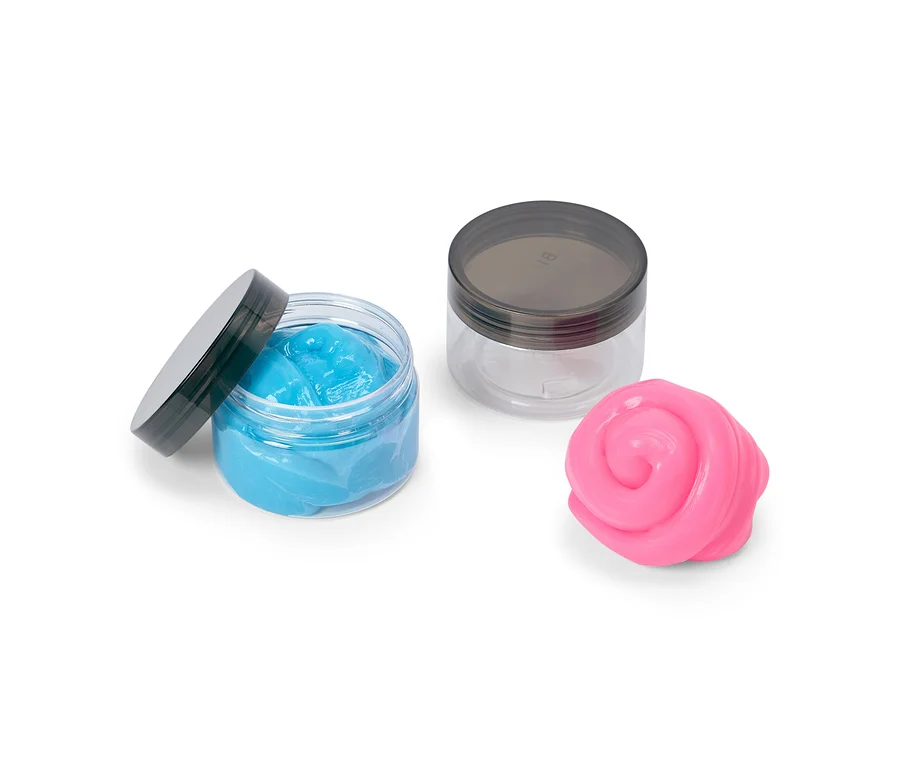 Zwei durchsichtige Dosen mit dunklen Deckeln; eine offene Dose enthält blauen Schleim, eine geschlossene Dose ist leer. Daneben liegt ein rosa Schleimklumpen.
