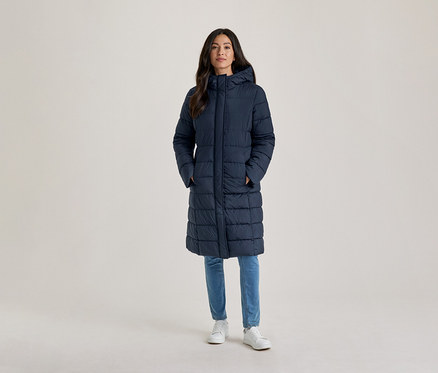 Frau steht, trägt einen langen, marineblauen, wattierten Mantel mit Kapuze, hellblaue Jeans und weiße Turnschuhe, die Hände in den Taschen.