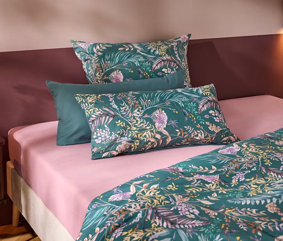 Ein Bett mit einem rosa Spannbettlaken und grüner Bettwäsche mit Blumen- und Blättermuster. Drei Kissen liegen am Kopfende, zwei gemustert und eines unifarben grün. Links steht ein Nachttisch mit einer weißen Vase und Blume vor einer zweifarbigen Wand.