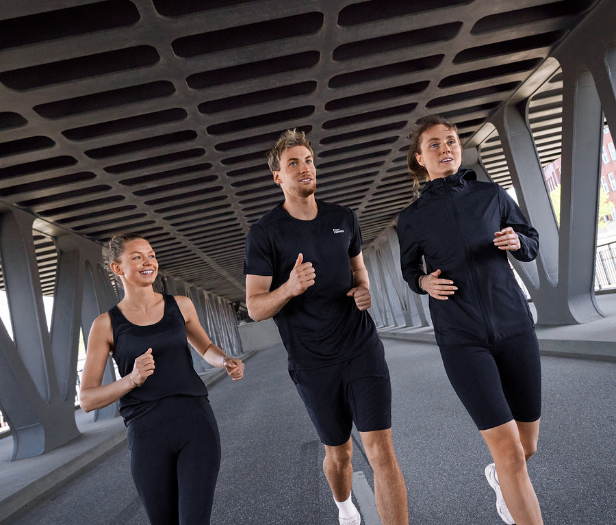 Drei Personen, zwei Frauen und ein Mann, laufen unter einer Brücke. Die Frau links trägt ein schwarzes Sport-Top und schwarze Sportleggings »ActivePower«. Der Mann in der Mitte trägt ein schwarzes Sportshirt und schwarze Sportradler »ActivePower«. Die Frau rechts trägt eine schwarze Windjacke »packable« und schwarze Sportradler »ActivePower«.