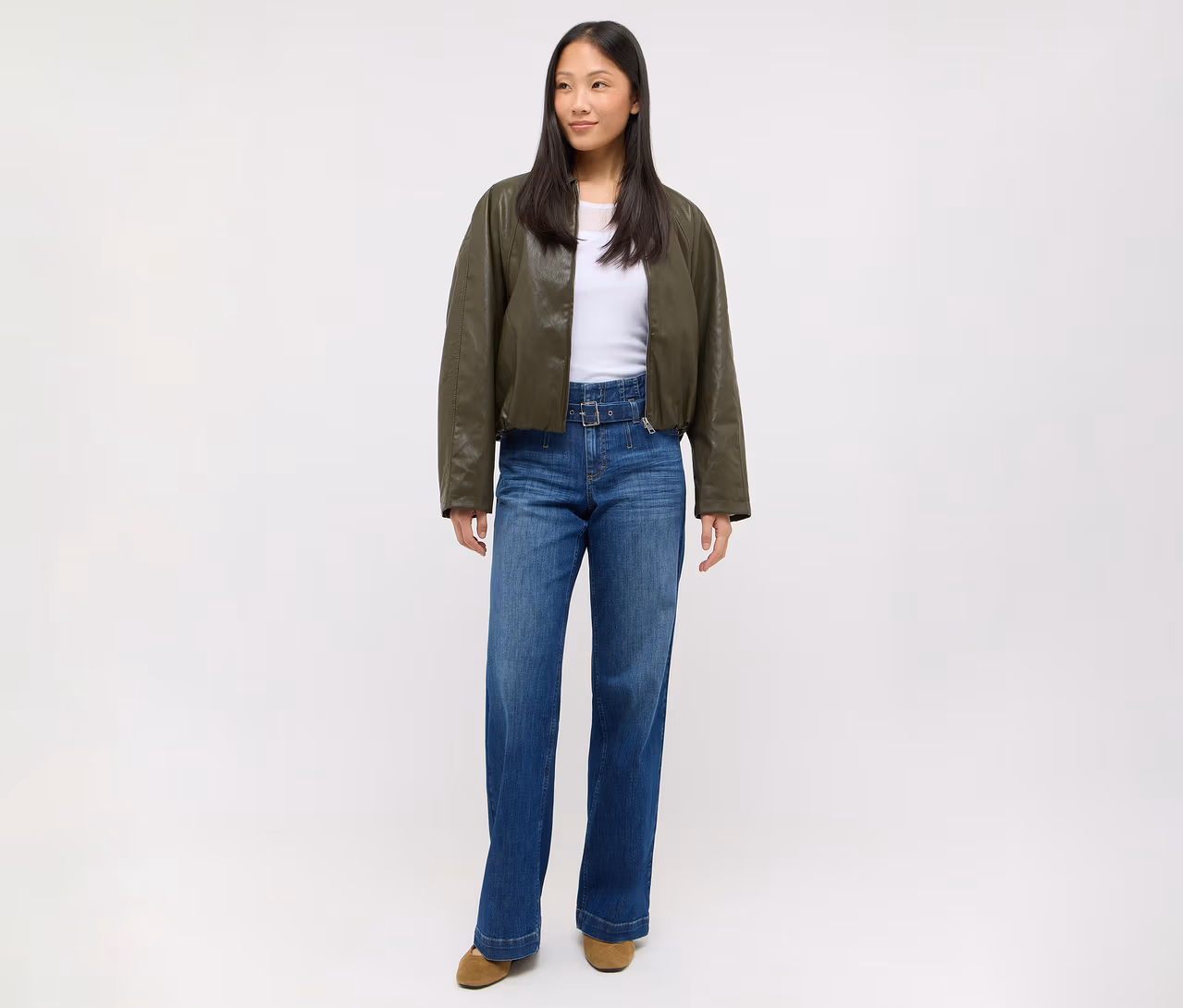 Frau steht, trägt eine dunkelgrüne Bomberjacke, ein weißes Oberteil, weite blaue Jeans mit Gürtel und hellbraune Schuhe.
