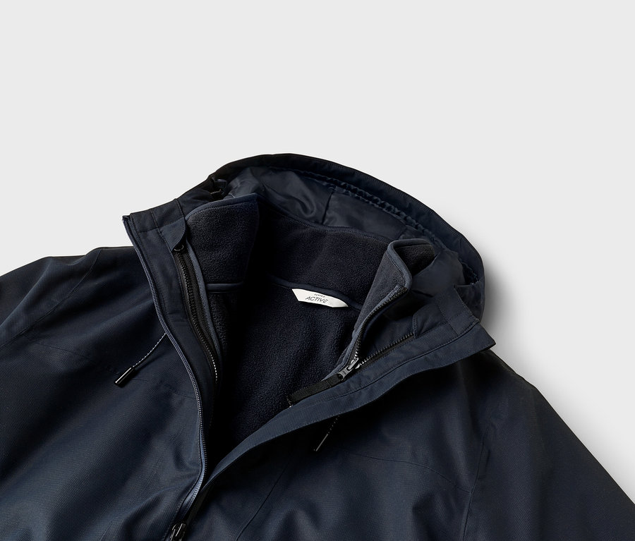 Nahaufnahme einer dunkelblauen Lagenjacke mit Außenhülle und Innenfleece. Die Jacke hat eine Kapuze, mehrere Reißverschlüsse und ein weißes „ACTIVE“-Etikett am Fleece.