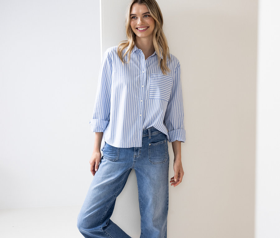 Eine lächelnde Frau mit langen blonden Haaren trägt eine blau-weiß gestreifte Street One Hemdbluse mit hochgekrempelten Ärmeln und eine Jeans, während sie an einer weißen Wand lehnt.
