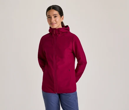 Frau in einer bordeauxroten Kapuzenjacke und blauen Hosen, die lächelt und die Hände hinter dem Rücken hält.