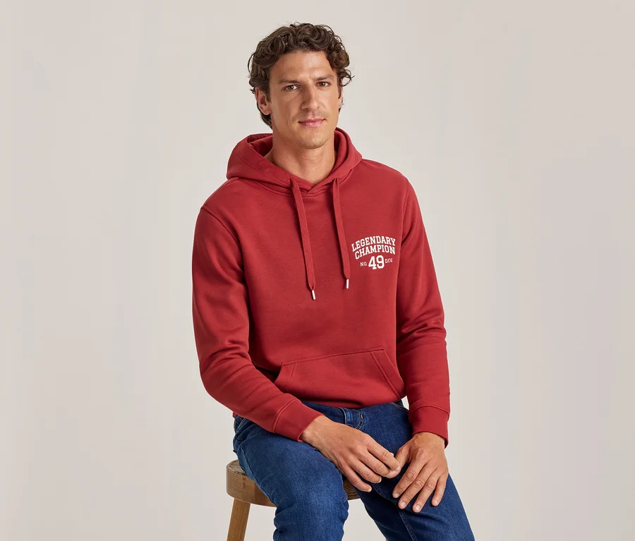 Mann mit lockigem Haar sitzt auf einem Hocker, trägt einen roten Hoodie mit „LEGENDARY CHAMPION No. 49 DIV“-Aufdruck und blaue Jeans.