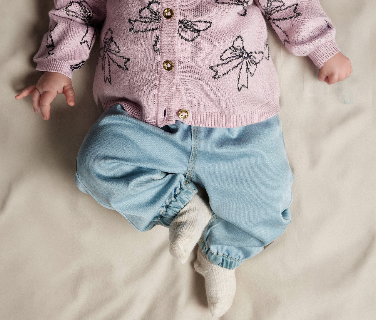 Baby liegt auf dem Rücken und trägt eine rosa Strickjacke mit schwarzen Schleifenmustern und goldenen Knöpfen, hellblaue Hosen und weiße Socken.
