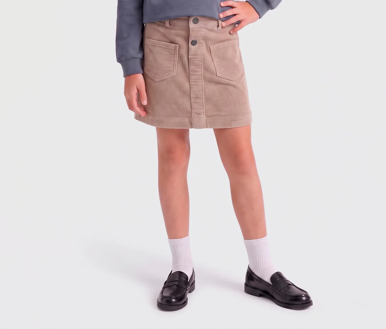 Kind trägt einen beigefarbenen Cord-Minirock mit Taschen und Knöpfen, ein graues Oberteil, weiße Socken und schwarze Loafer.