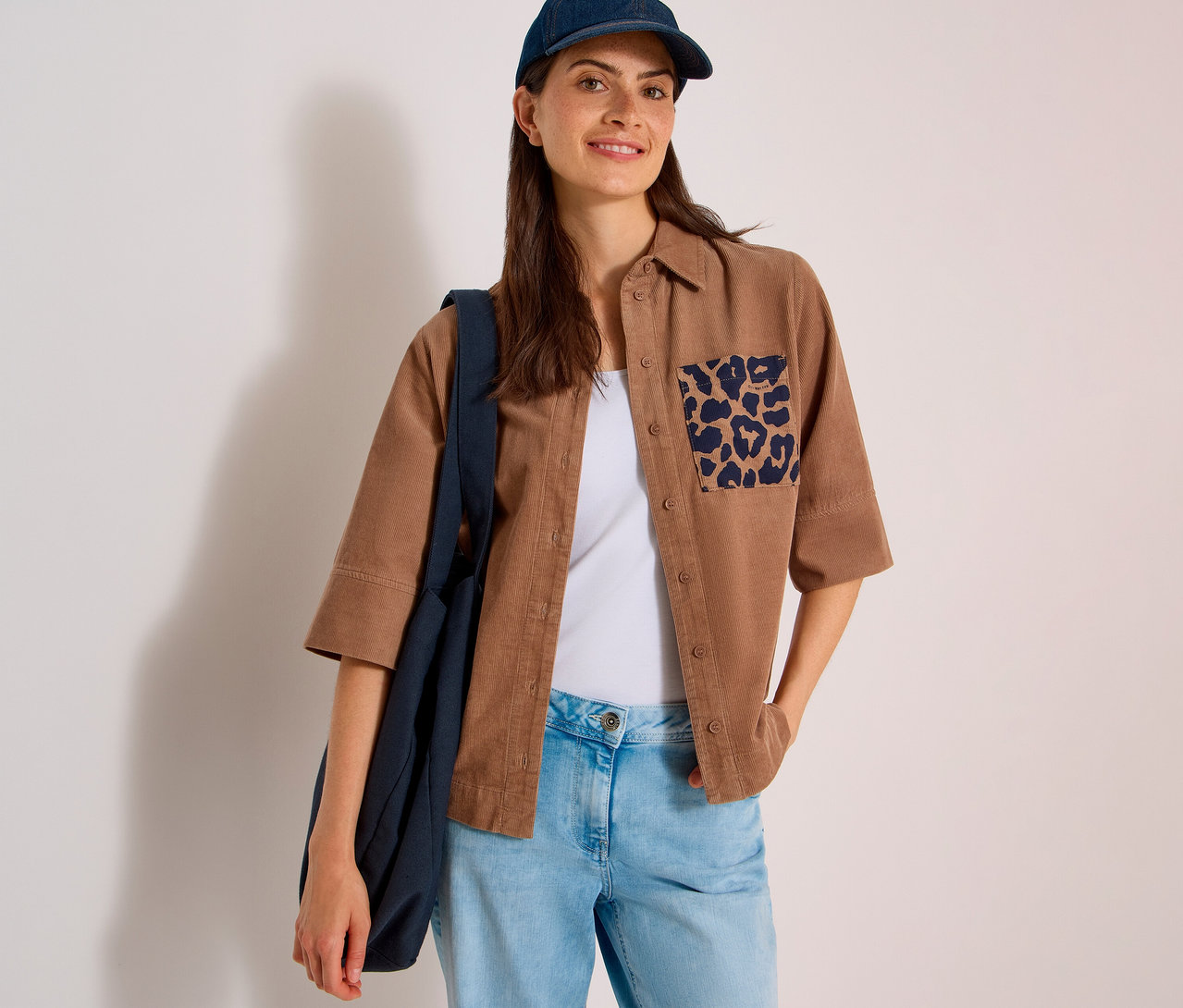 Lächelnde Frau mit dunkelblauer Kappe, trägt eine braune CECIL Hemdbluse mit Leopardenmuster-Tasche, helle Jeans und eine dunkelblaue Tasche.