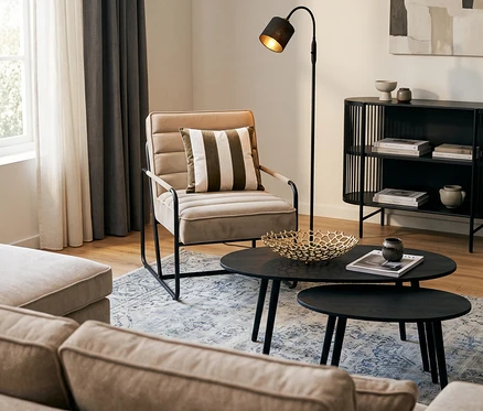 Ein Wohnzimmer mit einem beigen Ecksofa »Veluno«, einem cremefarbenen Eglo Stuhl »Pakotkuara« mit gestreiftem Kissen, zwei schwarzen Eglo Tischen »ALORAN« und einer schwarzen Reality Stehleuchte »Tommy«. Ein Benuta Teppich »Frencie« liegt am Boden, und ein grauer SCHÖNER WOHNEN-Kollektion Verdunkelungsvorhang »OPACO« hängt am Fenster. Im Hintergrund steht ein schwarzes Eglo Regal »DINSOR« und auf dem Tisch eine Schale »Kimitsu«.
