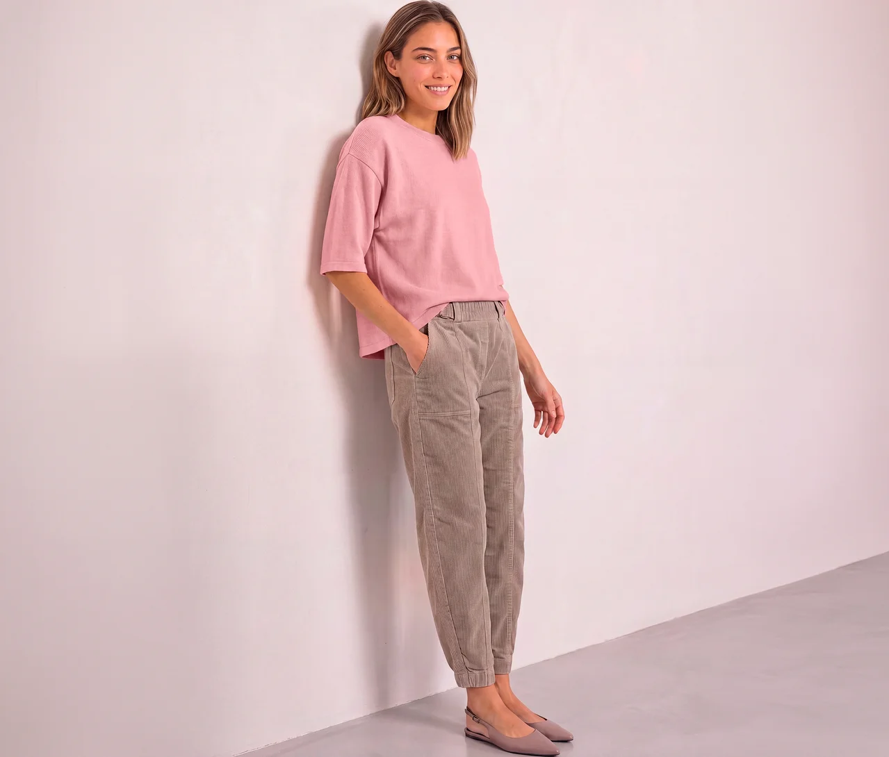 Lächelnde Frau lehnt an einer rosa Wand und trägt einen rosa Street One 3/4-Strickpullover, hellbraune Cordhose und hellrosa Ballerinas.